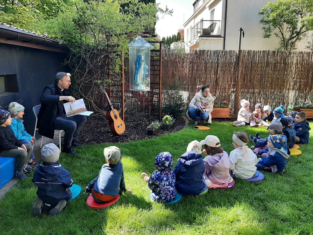 Kilkoro dzieci siedzi na trawie w ogrodzie. Siedzi tez ksiądz. Dzieci słuchają księdza. Dzieci modlą się przy figurze Maryi.